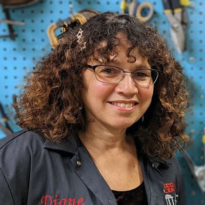 Smiling woman with curly brown hair and glasses, wearing a dark work shirt with the name 'Diane' embroidered on it, standing in front of a blue pegboard wall with tools.